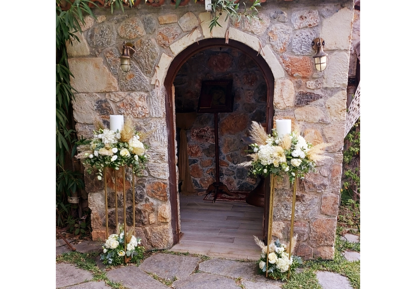 Λαμπάδες γάμου με λευκά άνθη, pampas grass και πρασινάδα σε μεταλλικές βάσεις μπροστά από πέτρινο εκκλησάκι στο Κτήμα Καλυψώ